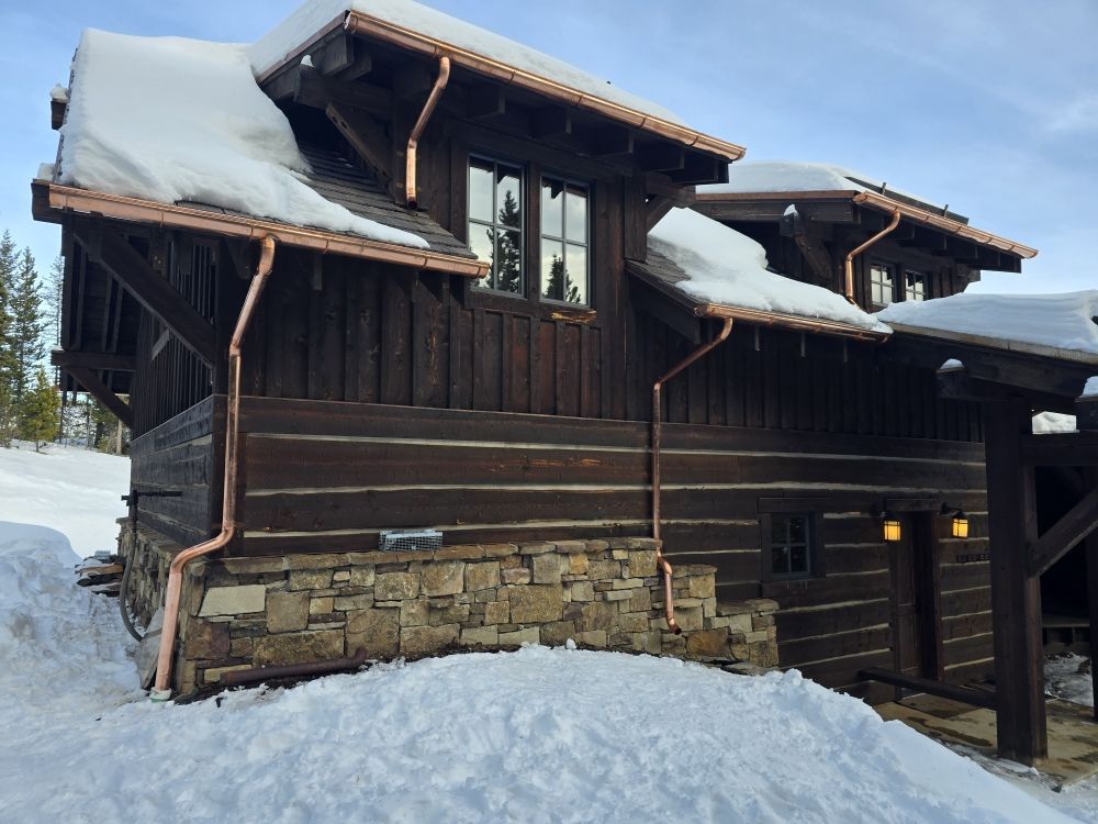 A rustic, dark-stained log cabin with a stone foundation and copper gutters, partially covered in snow during winter.