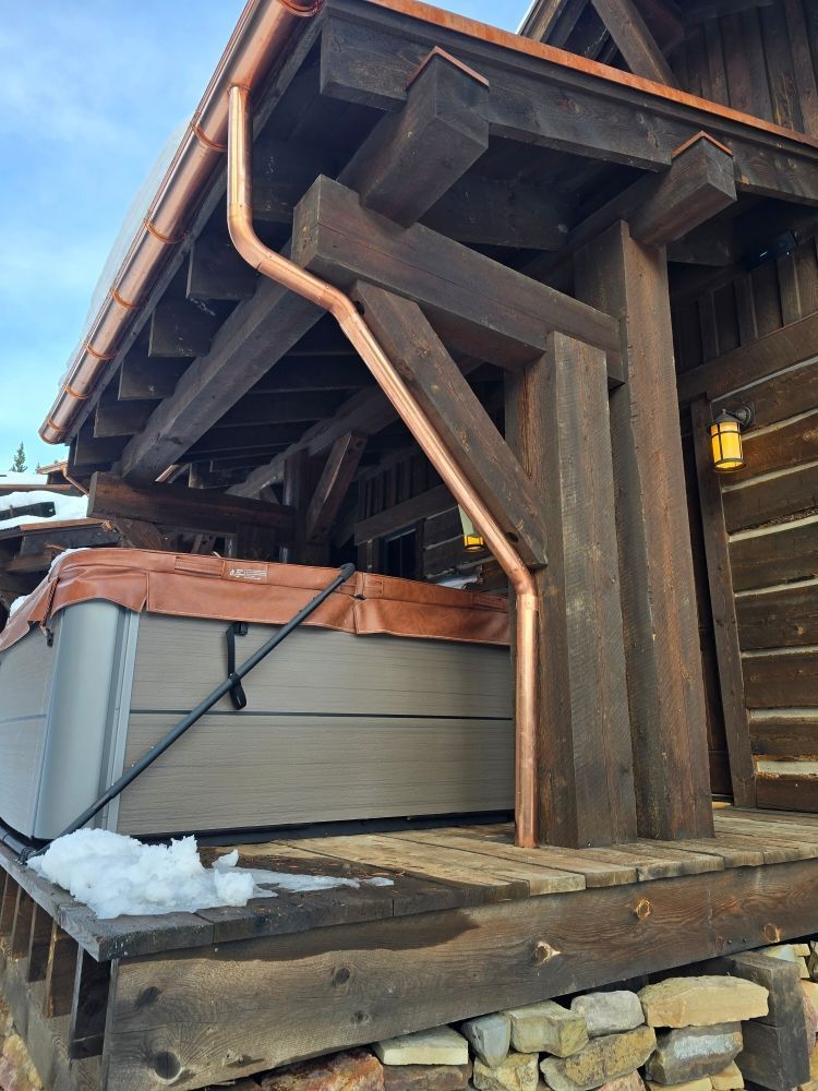 A copper downspout runs along a dark wooden timber post on a cabin porch, next to a hot tub cover.