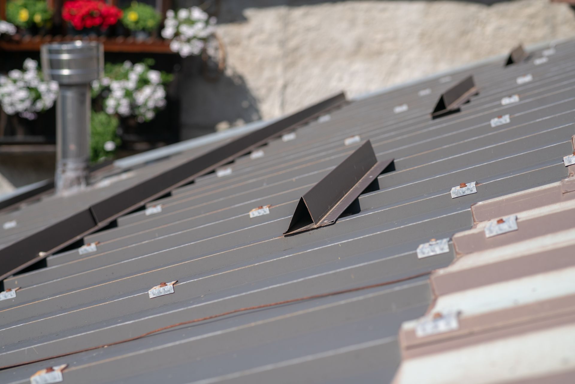 Dark metal roofing panels featuring several brown triangular snow guards and a metal chimney pipe near a floral display.