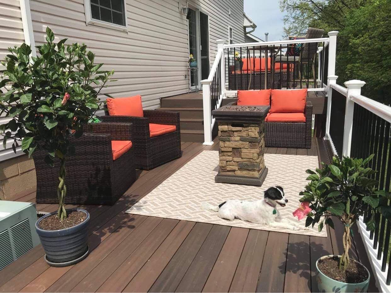 Patio with seating, fire pit, dog, and potted plants. Brown deck, orange cushions, white and black railing.