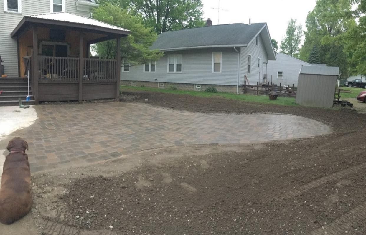 Dog sits on a partially paved patio with a house and another house in the background.