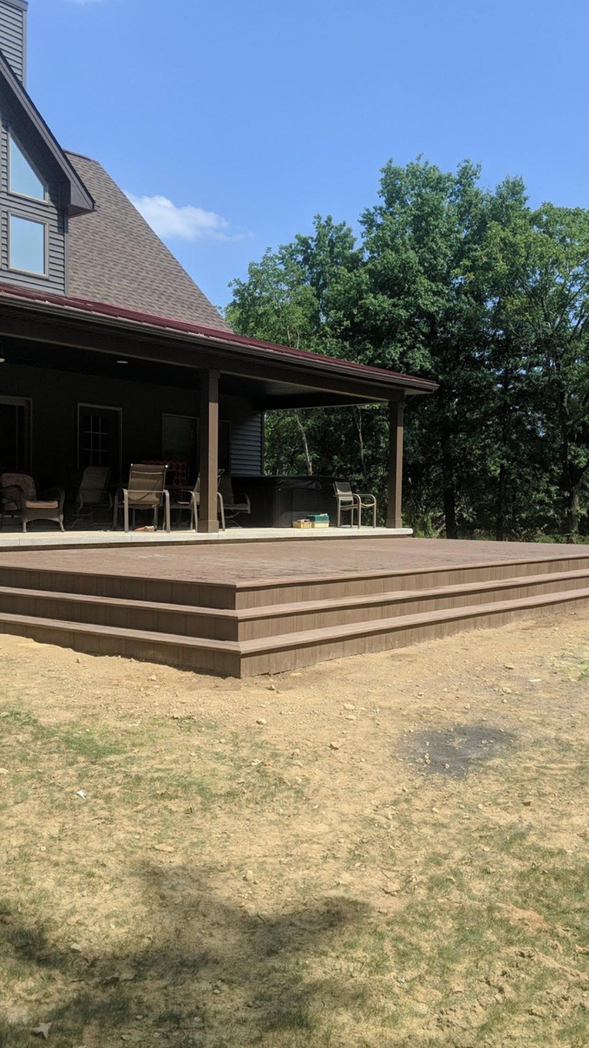 Brown wooden deck with steps leading to a house with pergola; green trees and blue sky.