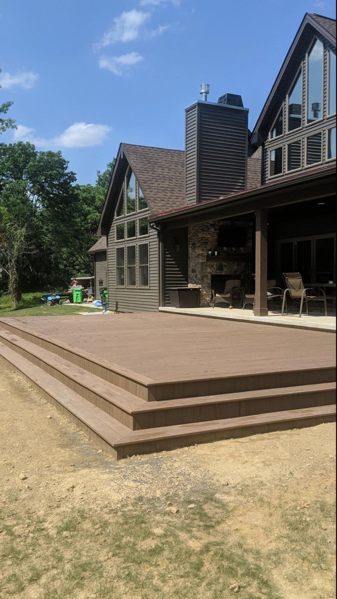 Brown deck and steps lead to a large house with a stone chimney and large windows on a sunny day.