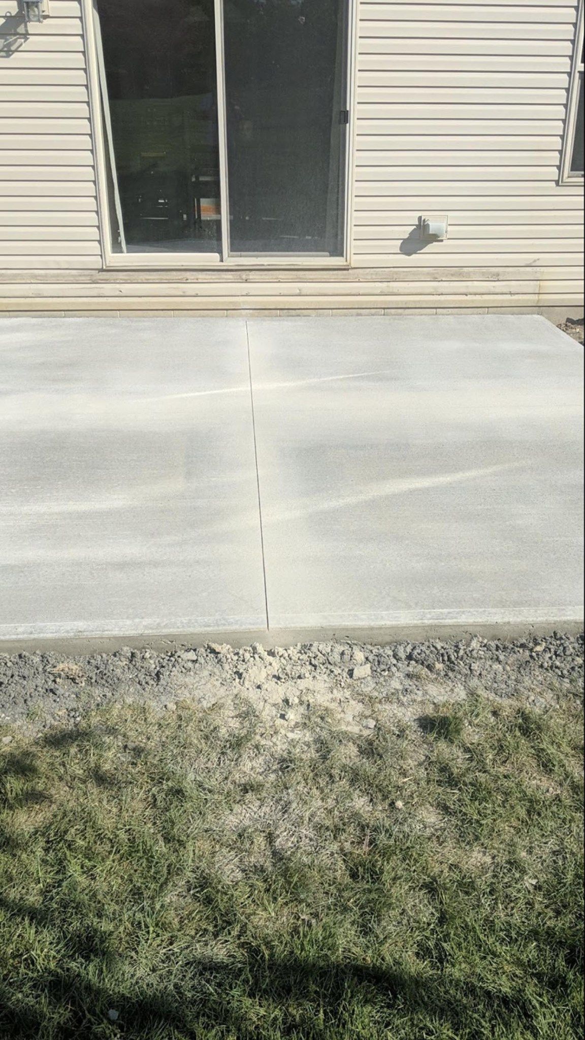 Concrete patio next to a house with a sliding glass door, edged with gravel and grass.