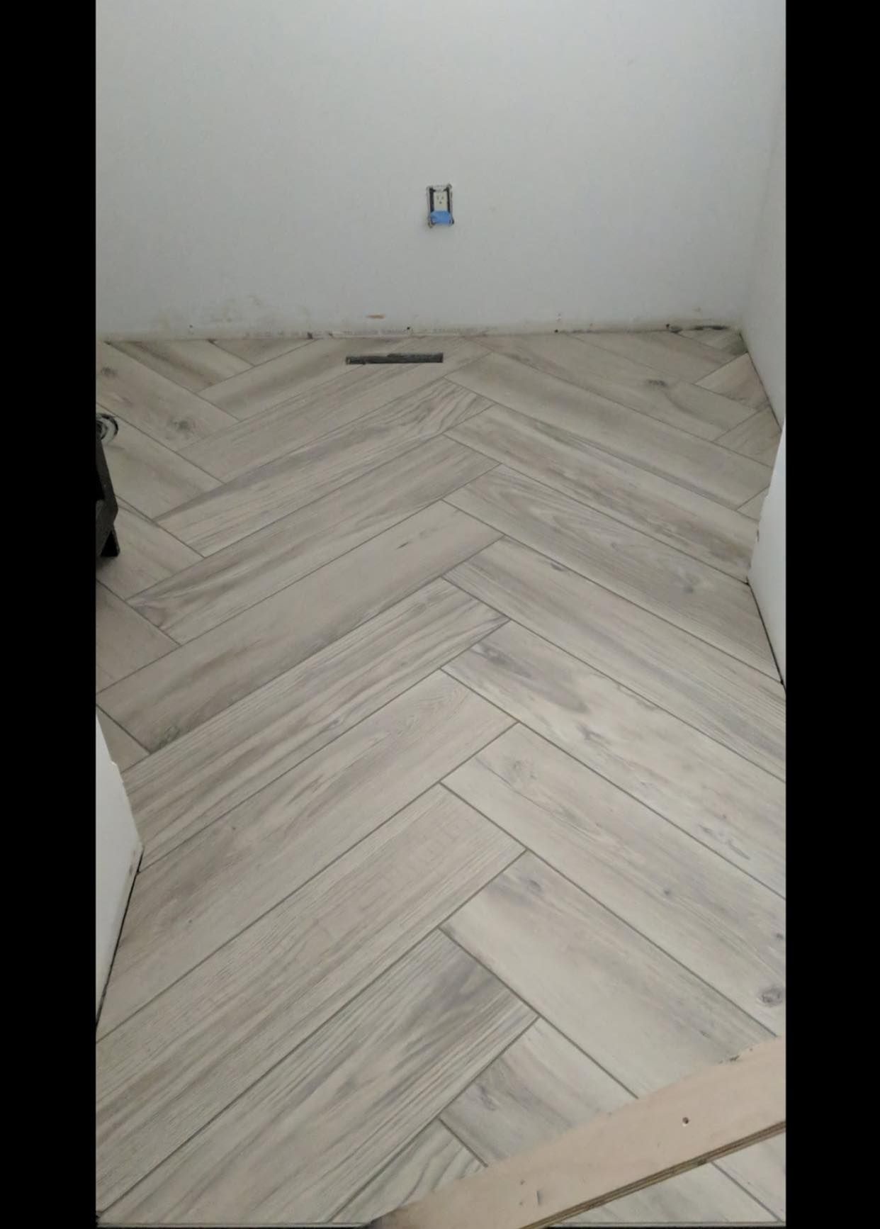 Herringbone pattern floor tiling in a bathroom under construction. Beige tiles, white walls, and an electrical box.