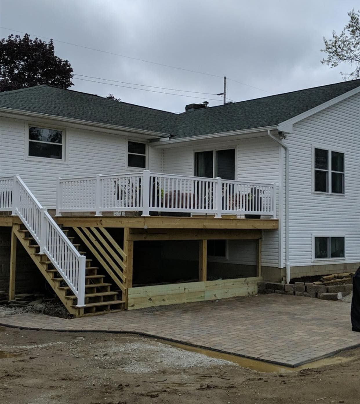 White house with a deck featuring stairs and white railings. A paved patio is below.