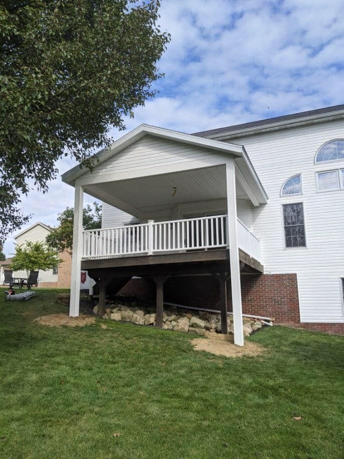 White-sided house with a covered deck, white railing, and wooden deck. Green grass and blue sky.