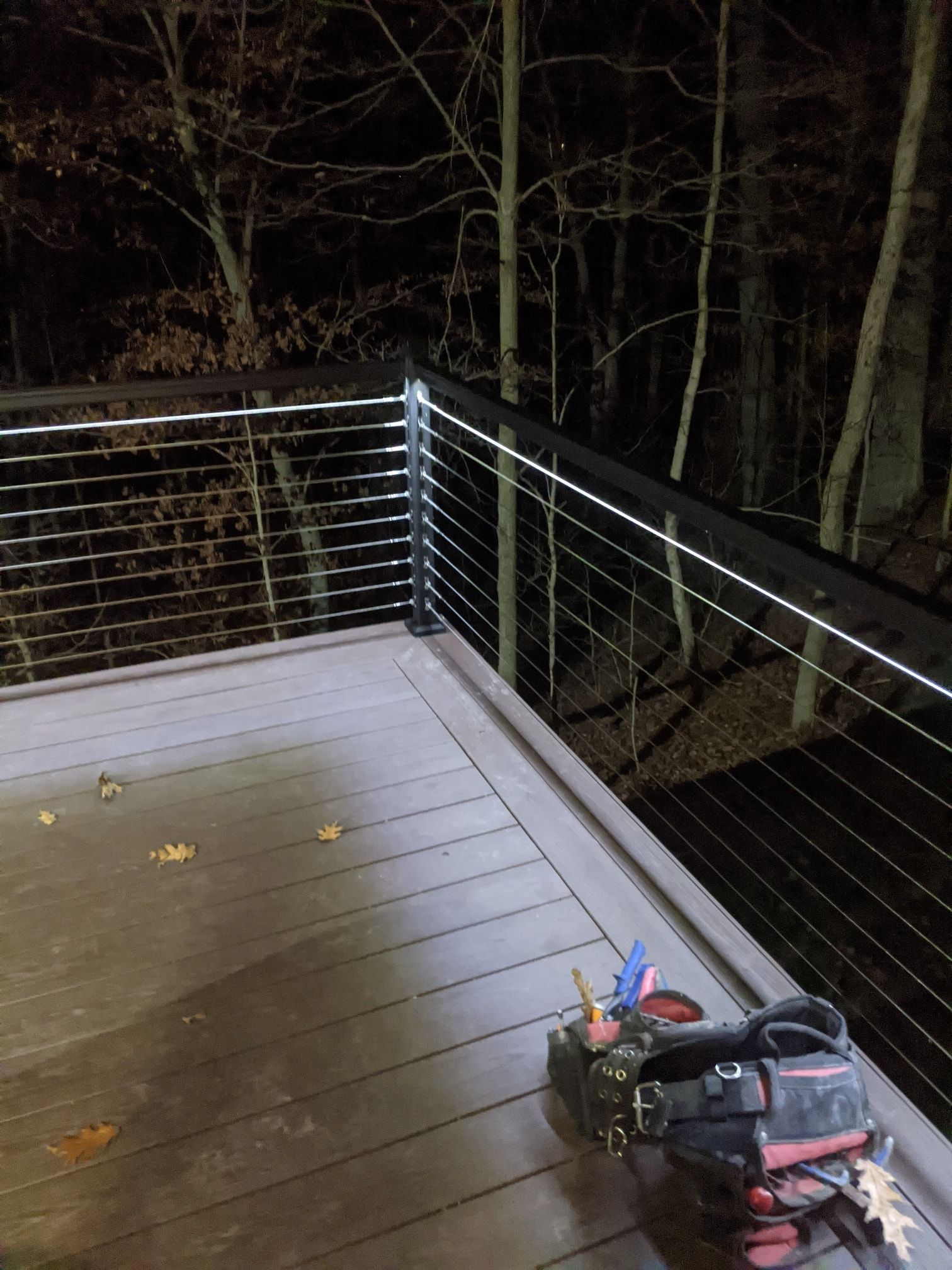Dark wooden deck with cable railing. Night setting with trees in the background and a toolbox on the deck.