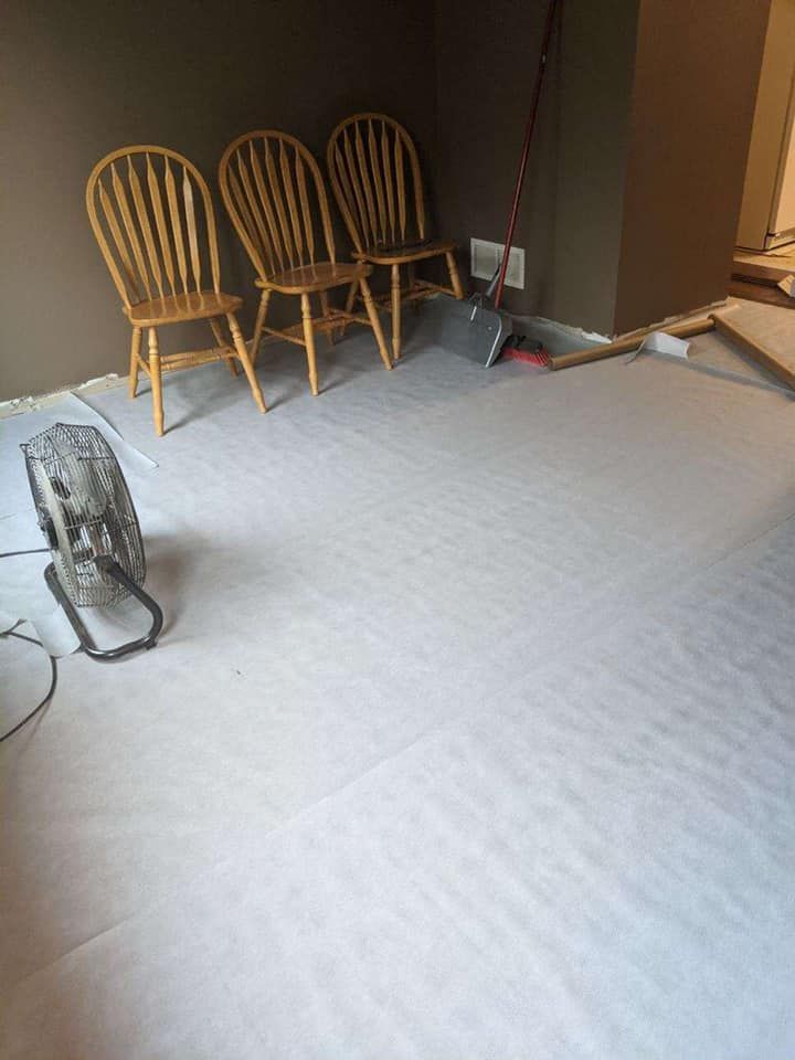 Three wooden chairs sit on flooring covered with white paper, a fan and broom are nearby.