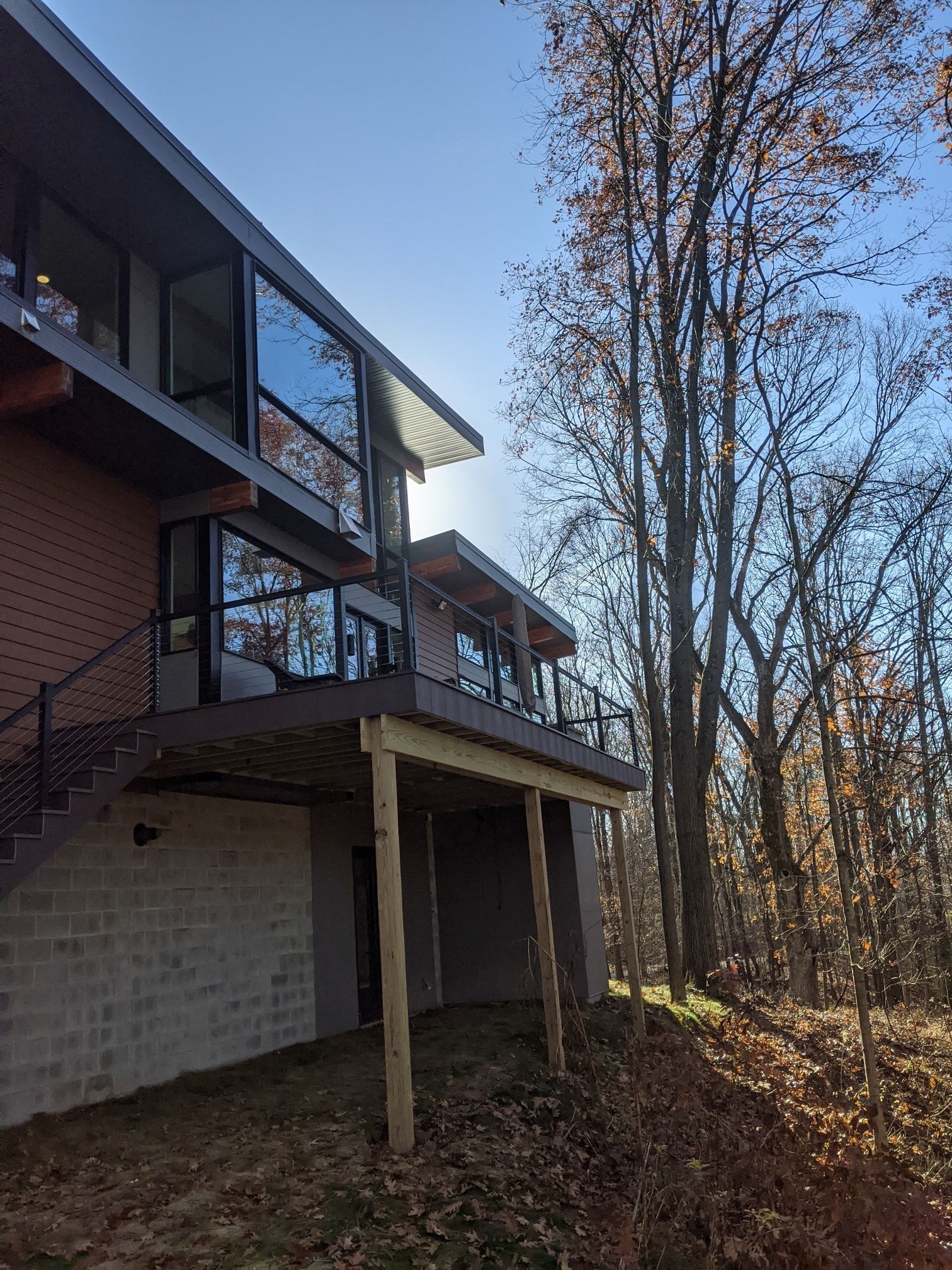 Modern two-story house with large windows, deck, and stairwell, set in a wooded area. Sunlight.