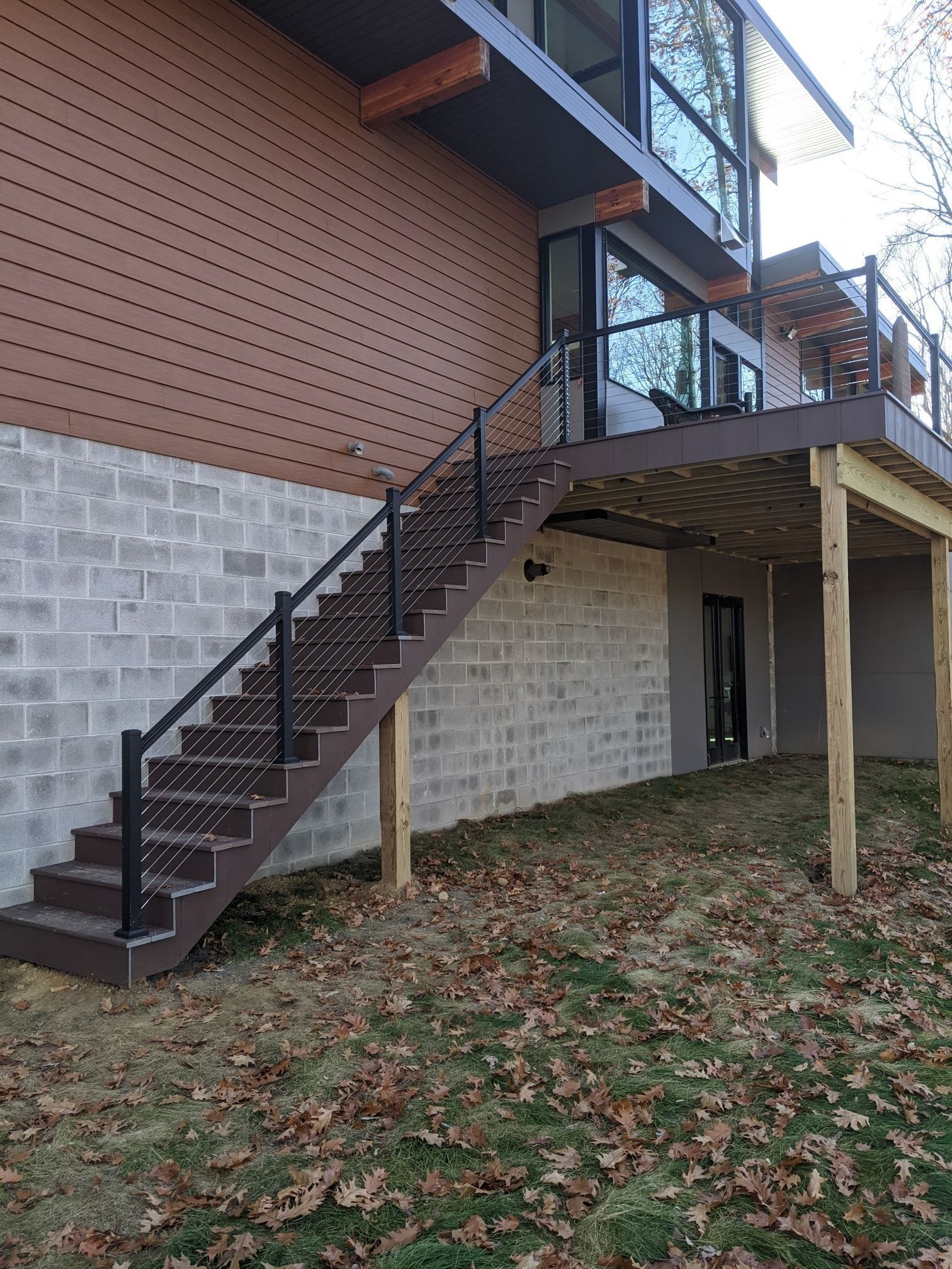 Brown stairs with black handrails lead to a deck attached to a brown and gray building with large windows.