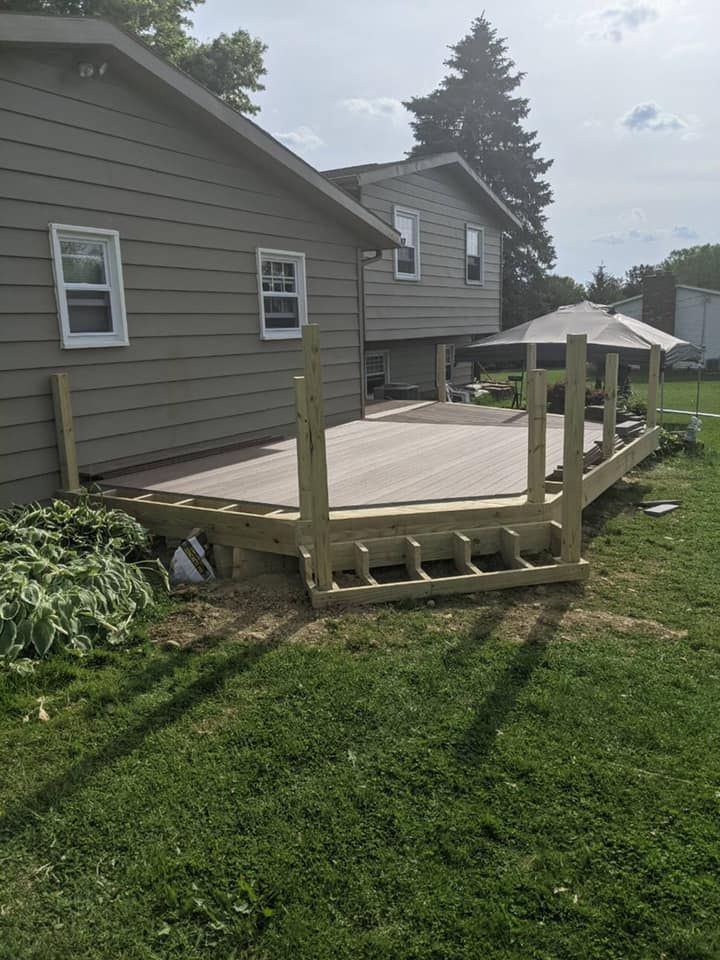 Wooden deck attached to a two-story house, surrounded by green grass and plants.