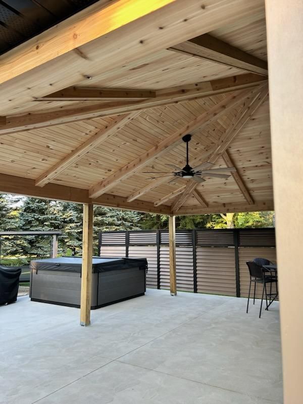 Patio with wooden gazebo, hot tub, and ceiling fan. Grey concrete floor, trees in the background.