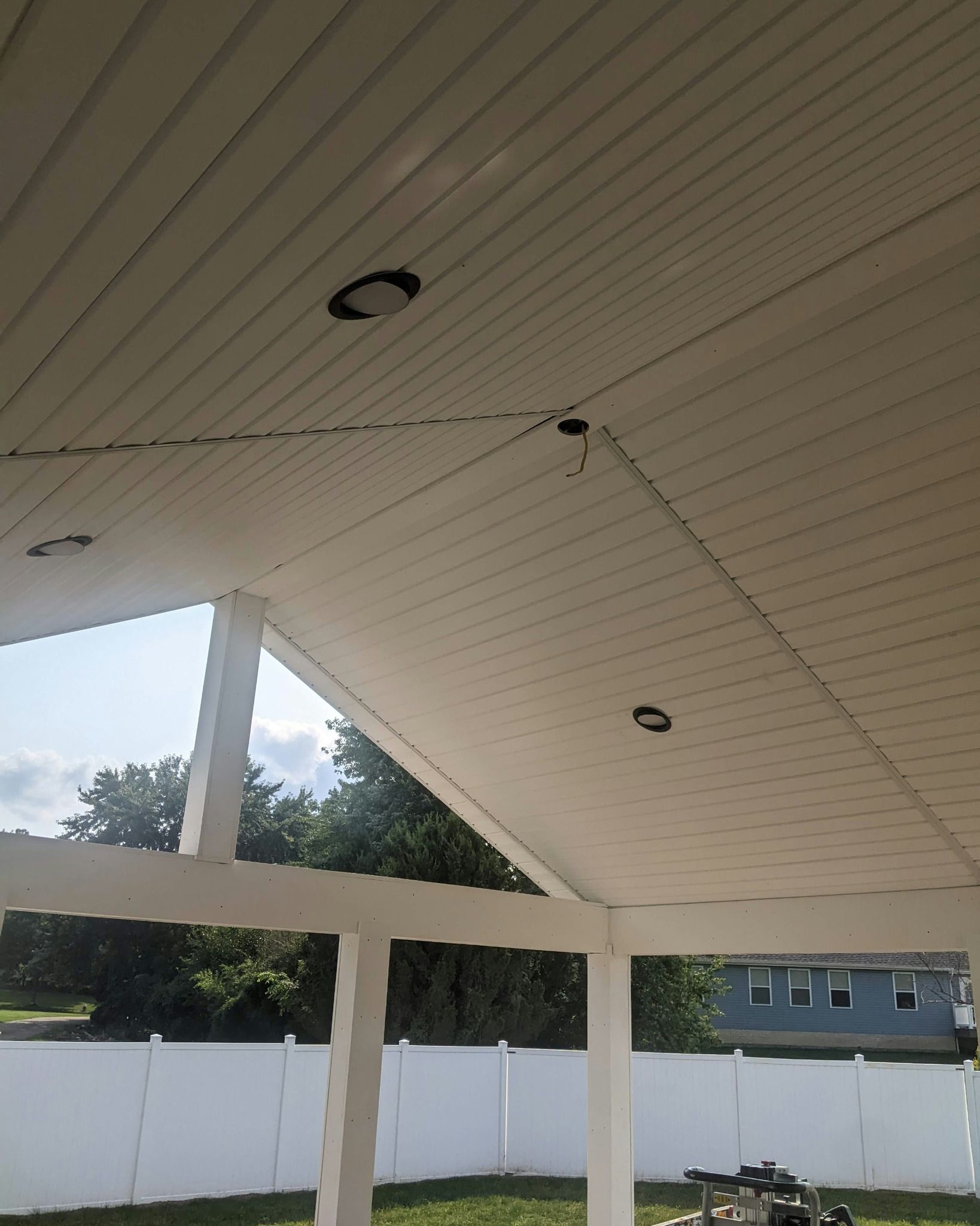 White outdoor ceiling with recessed lights, supported by white posts, with trees and a fence in the background.