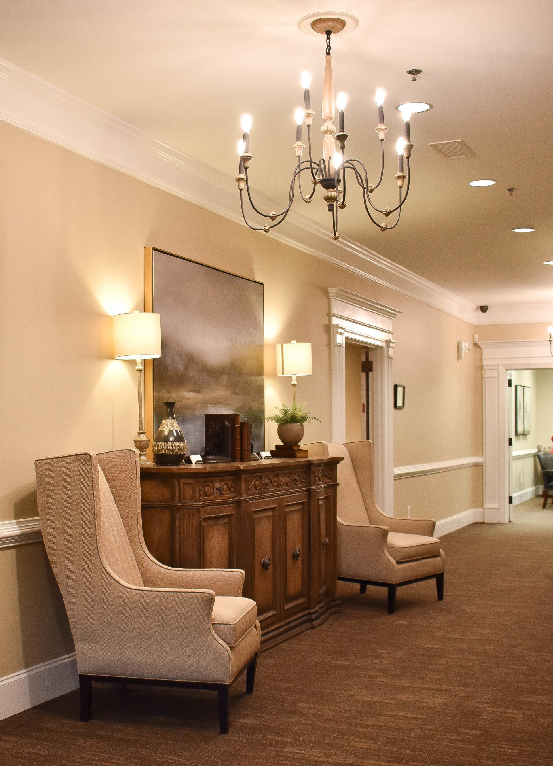 A hallway with chairs , a dresser and a chandelier.