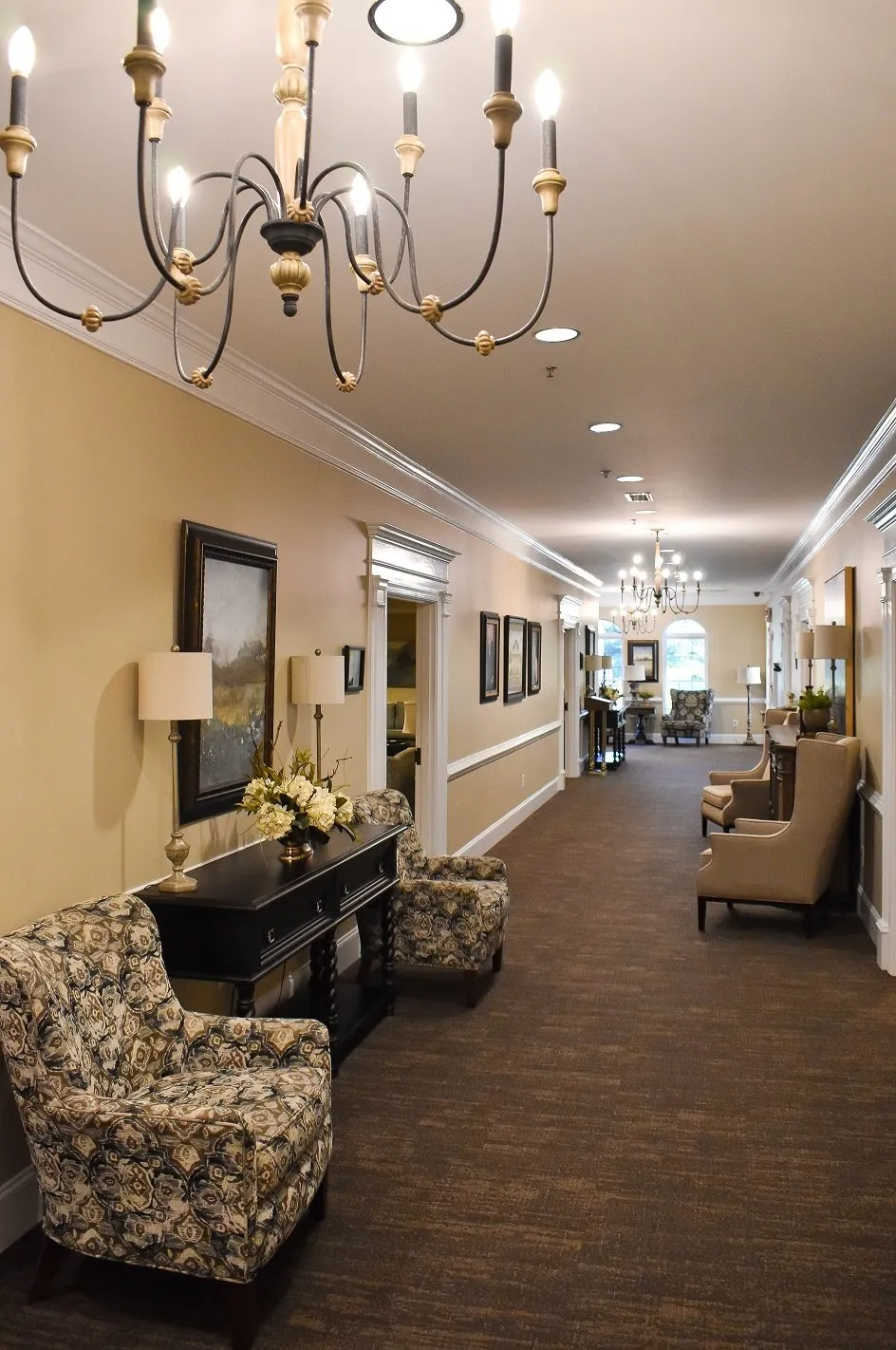 A long hallway with chairs , a table and a chandelier hanging from the ceiling.