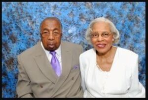 Elderly couple in formal attire posing for a photo against a blue mottled backdrop.