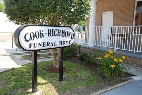 Sign for Cook-Richmond Funeral Home; black and white oval sign, two black posts, green lawn and flowers.
