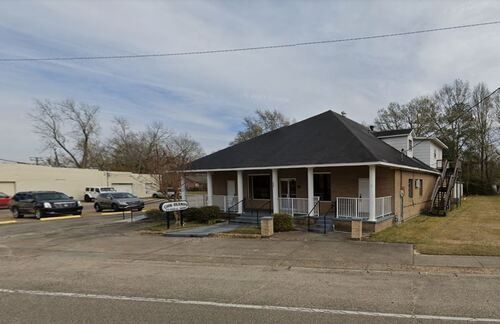 A brick building with a dark roof and a porch. Cars parked in front on a paved lot.