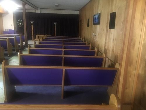 Rows of purple-padded pews in a chapel. Wooden walls and flooring. Dark curtain in the background.