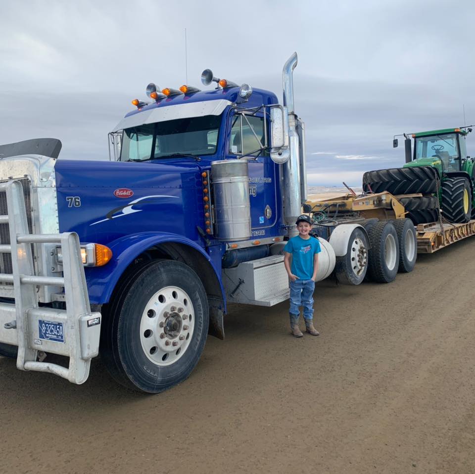A person stands next to a blue semi-truck and trailer hauling a green tractor on a dirt road.