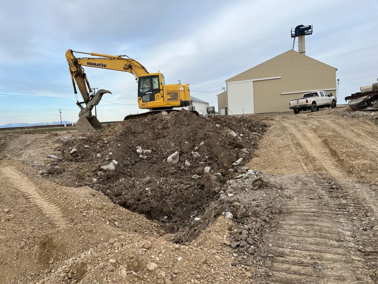 Yellow excavator digging into a mound of dirt near a beige building and a white truck.