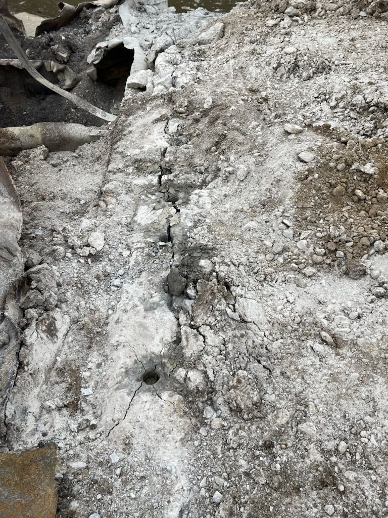 Ground surface with a trench dug down the center. Light gray and brown debris covers the area.