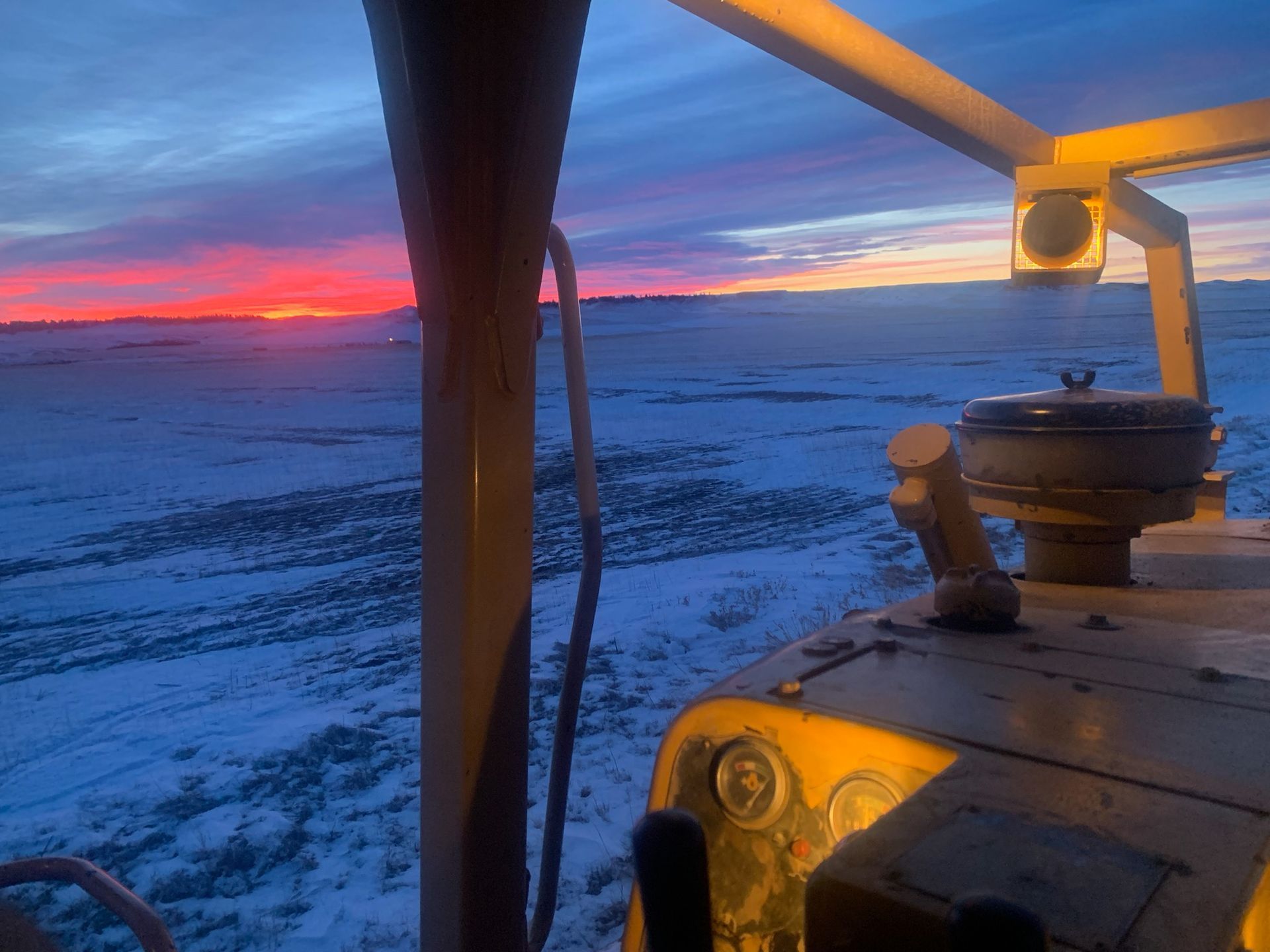 Sunrise over a snowy field seen from inside a yellow tractor cab.