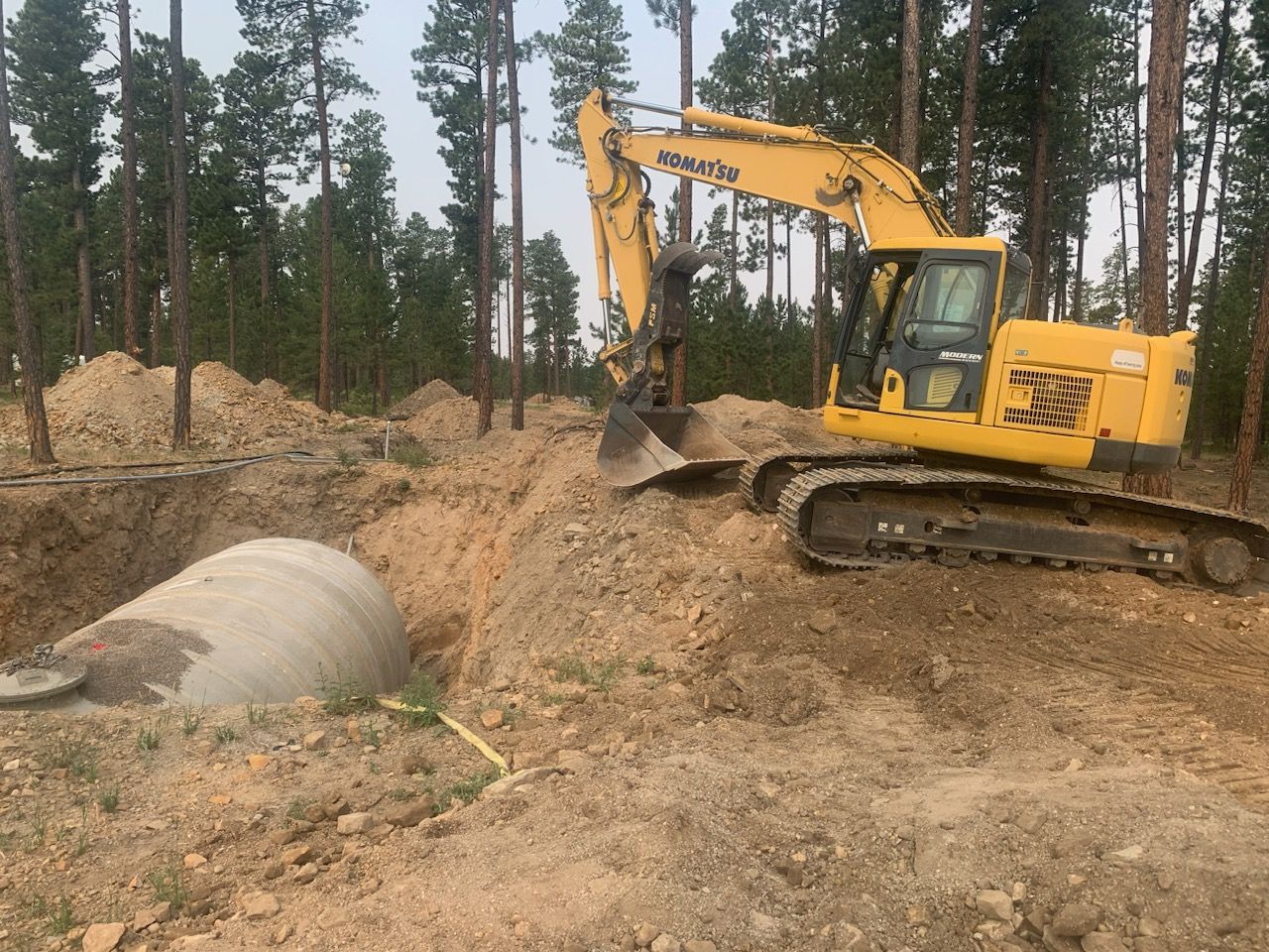 Yellow excavator digs around a large concrete culvert in a forest setting.