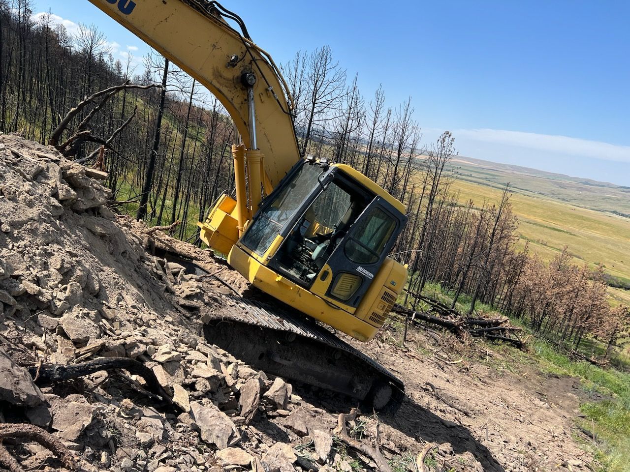 Yellow excavator on a steep hillside, digging into the earth; partially burned trees in the background.