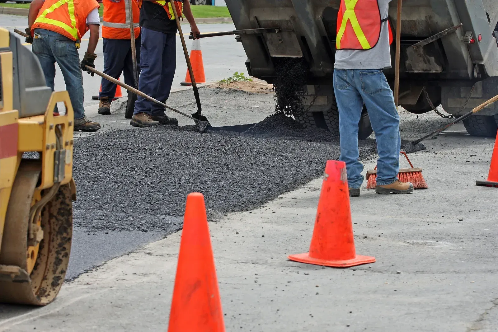 Road workers in orange vests paving a street with asphalt, orange cones nearby.