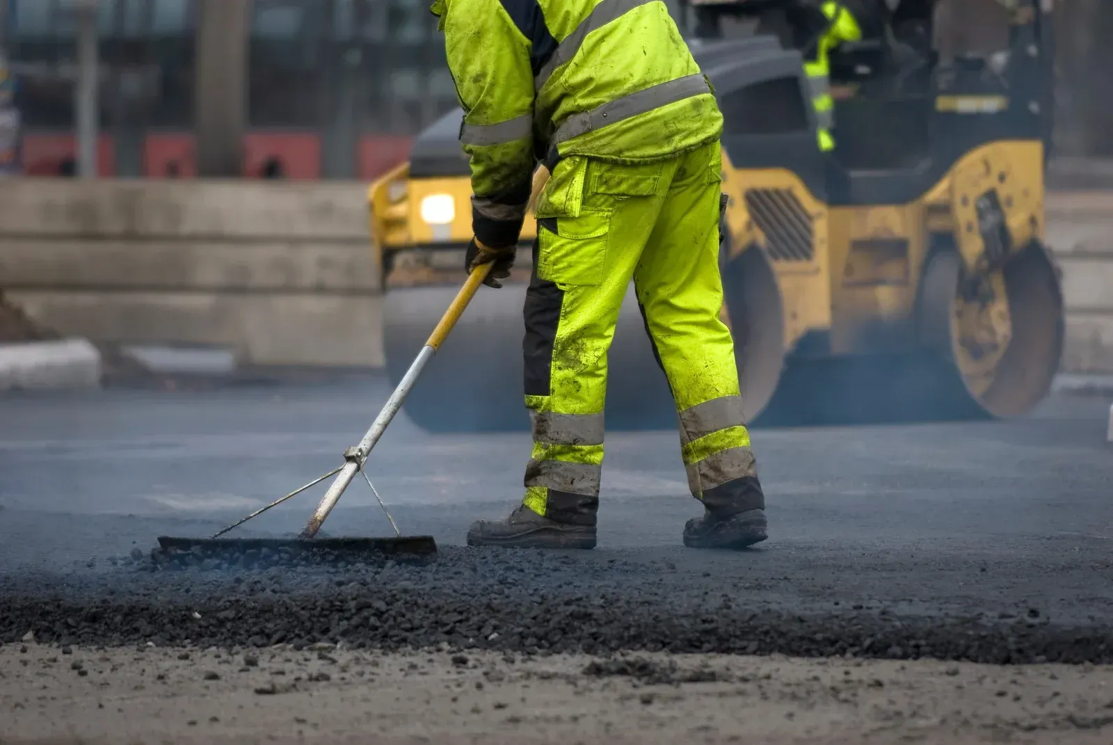 Road worker in yellow reflective suit using a rake to spread asphalt. Roller in the background.