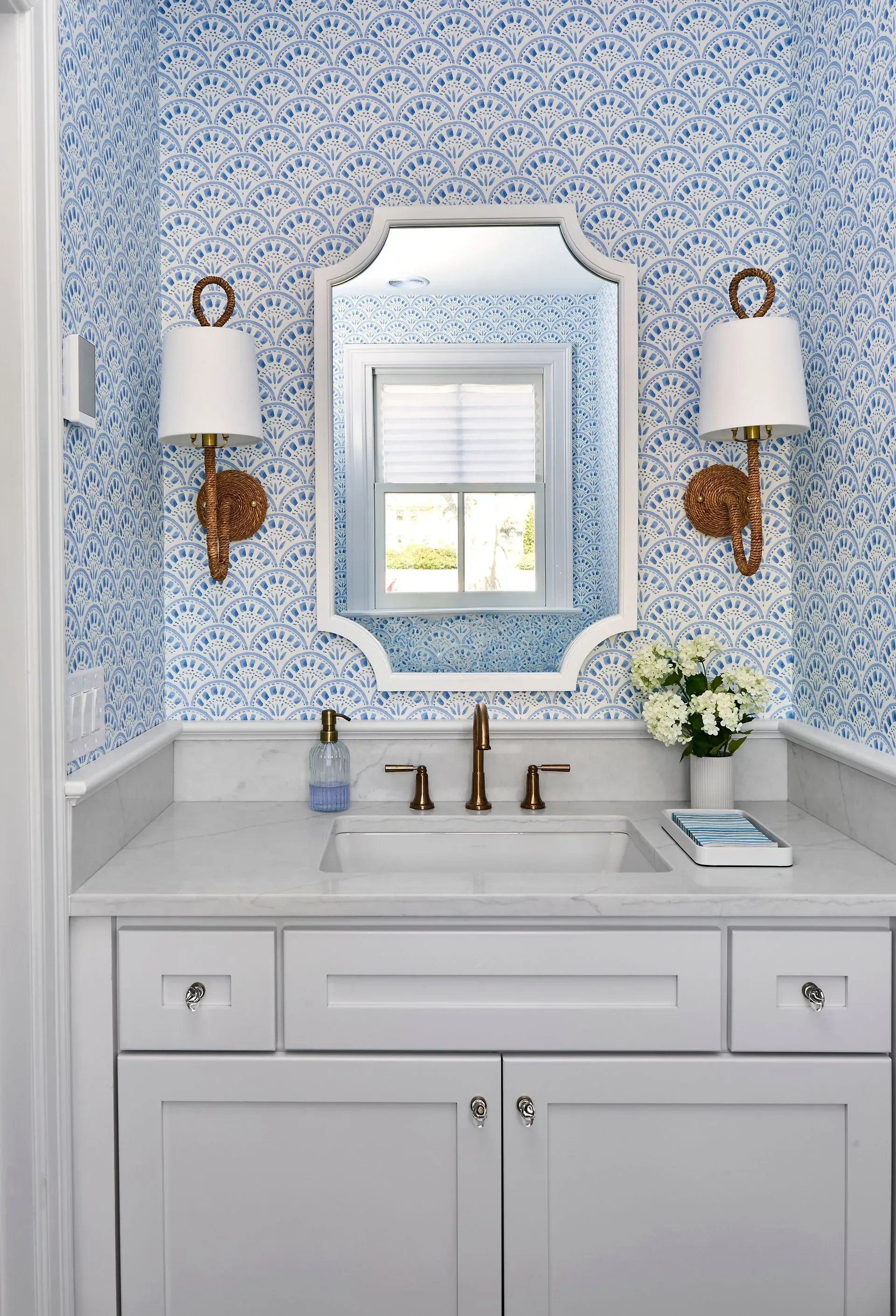 White vanity with blue patterned wallpaper, framed mirror, sconces, and sink.