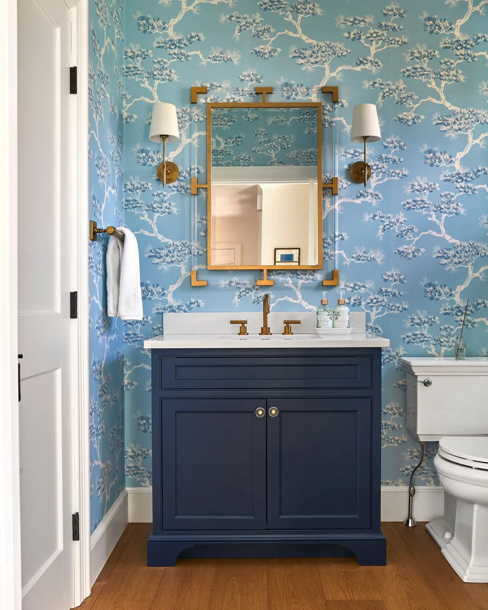 Blue and white floral wallpapered powder room with navy vanity, gold mirror, and white toilet.