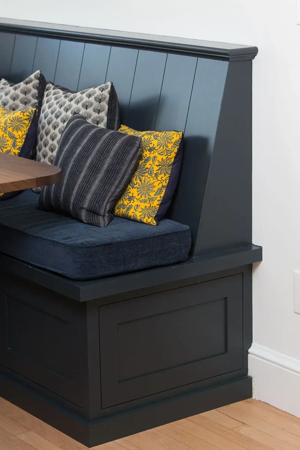 Built-in navy blue dining bench with cushions and pillows, near a table. White wall, wooden floor.