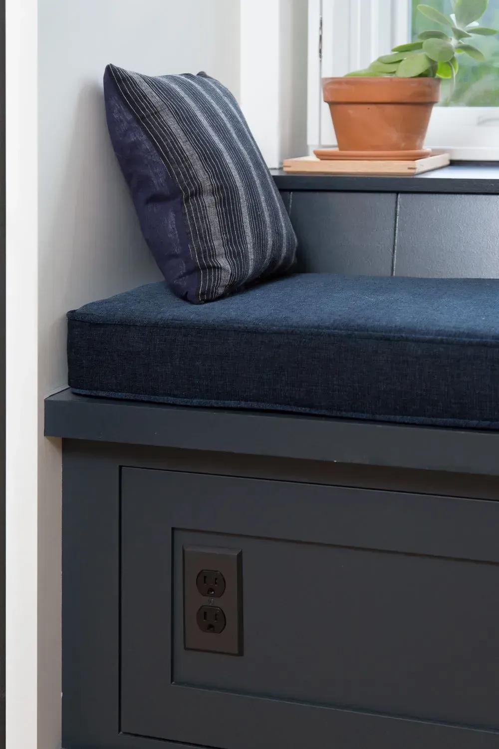 Built-in navy blue window seat with cushion and decorative pillow; brown outlet; terracotta pot with plant on windowsill.