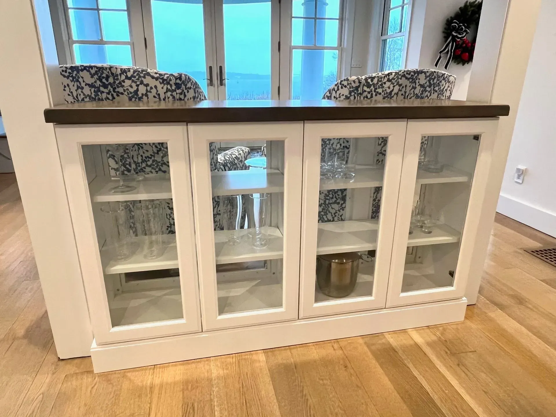 White cabinet with glass doors and wooden countertop. Contains glassware, set against a window view.