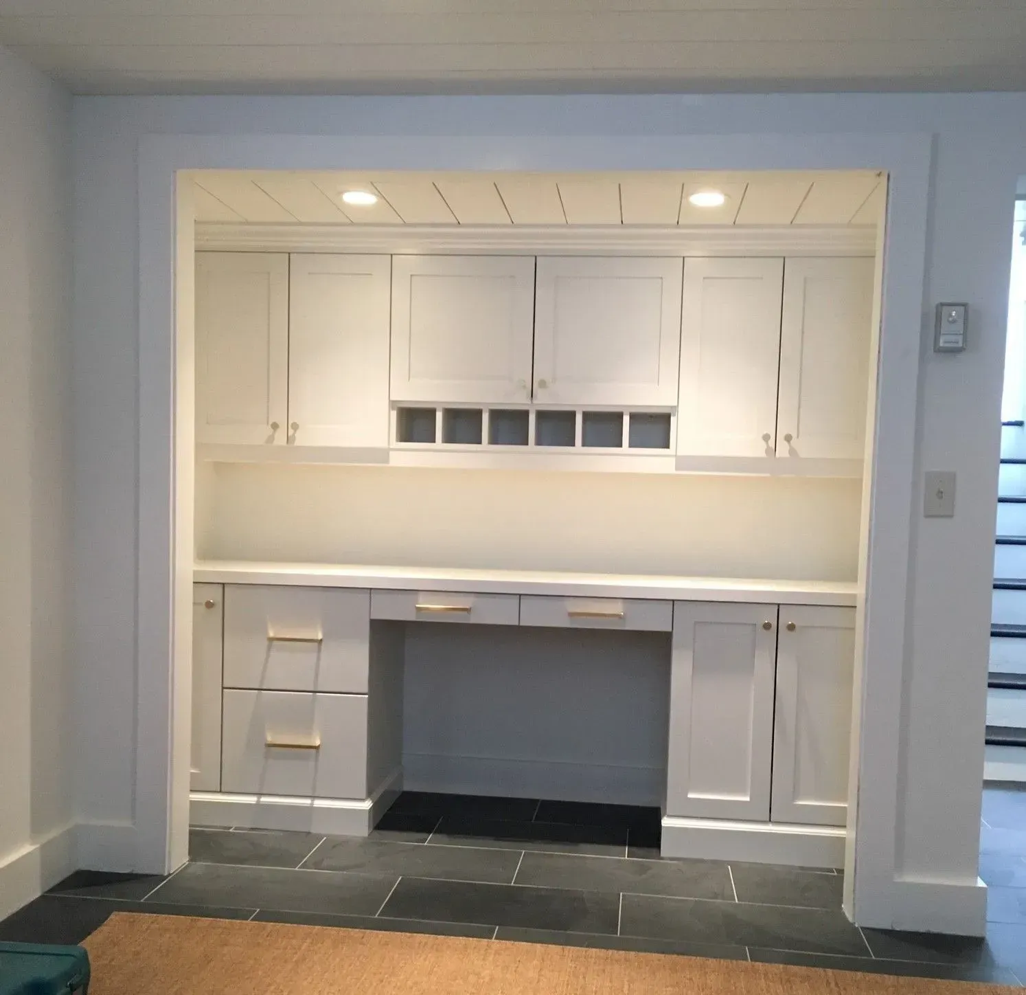 Built-in white desk with cabinets, lights, and wine rack, in an alcove with black tile floor.