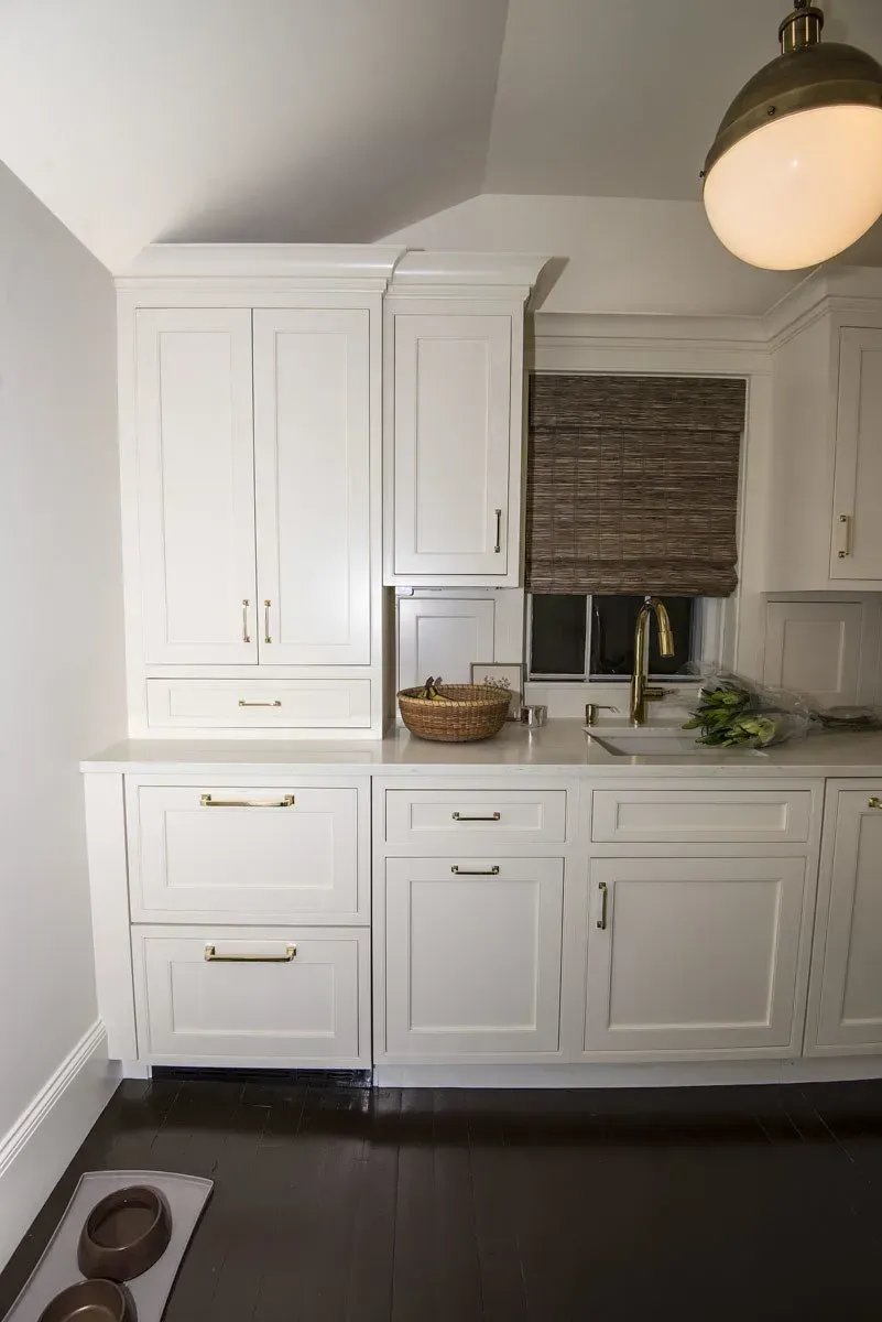 White kitchen cabinets with gold hardware, dark floors, and a woven window shade.