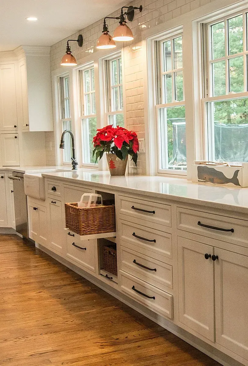 White kitchen with marble countertop, wood floor, and windows, featuring red poinsettias and black hardware.