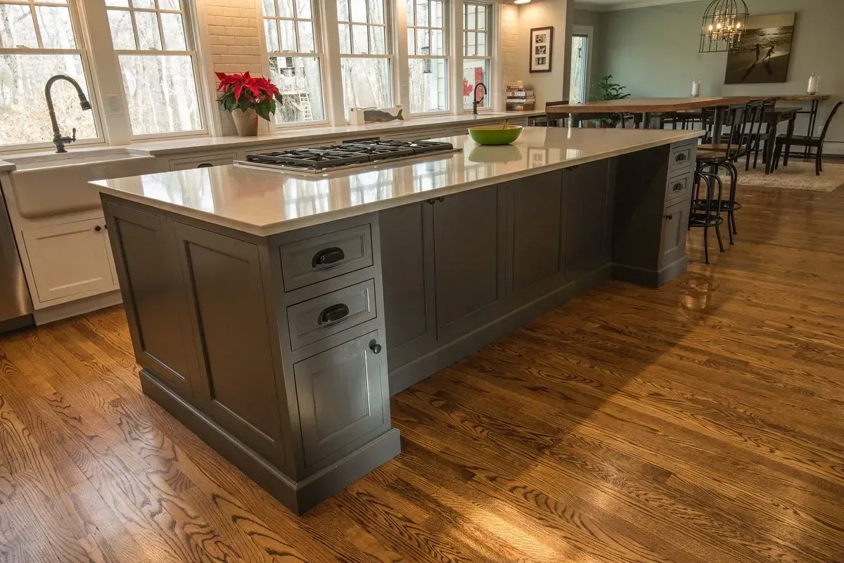 Gray kitchen island with cooktop, set in a light-filled kitchen with hardwood floors.
