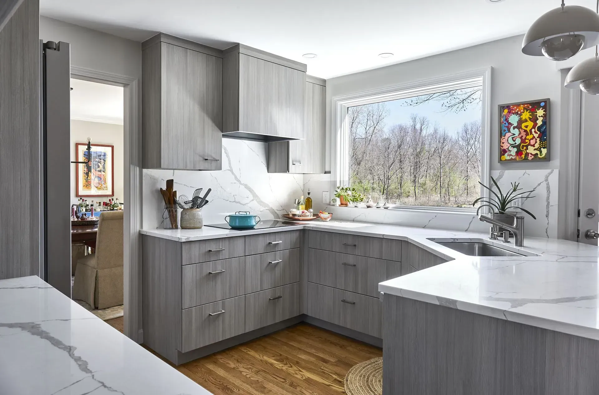 Modern kitchen with gray cabinets, marble countertops, and a window overlooking trees.