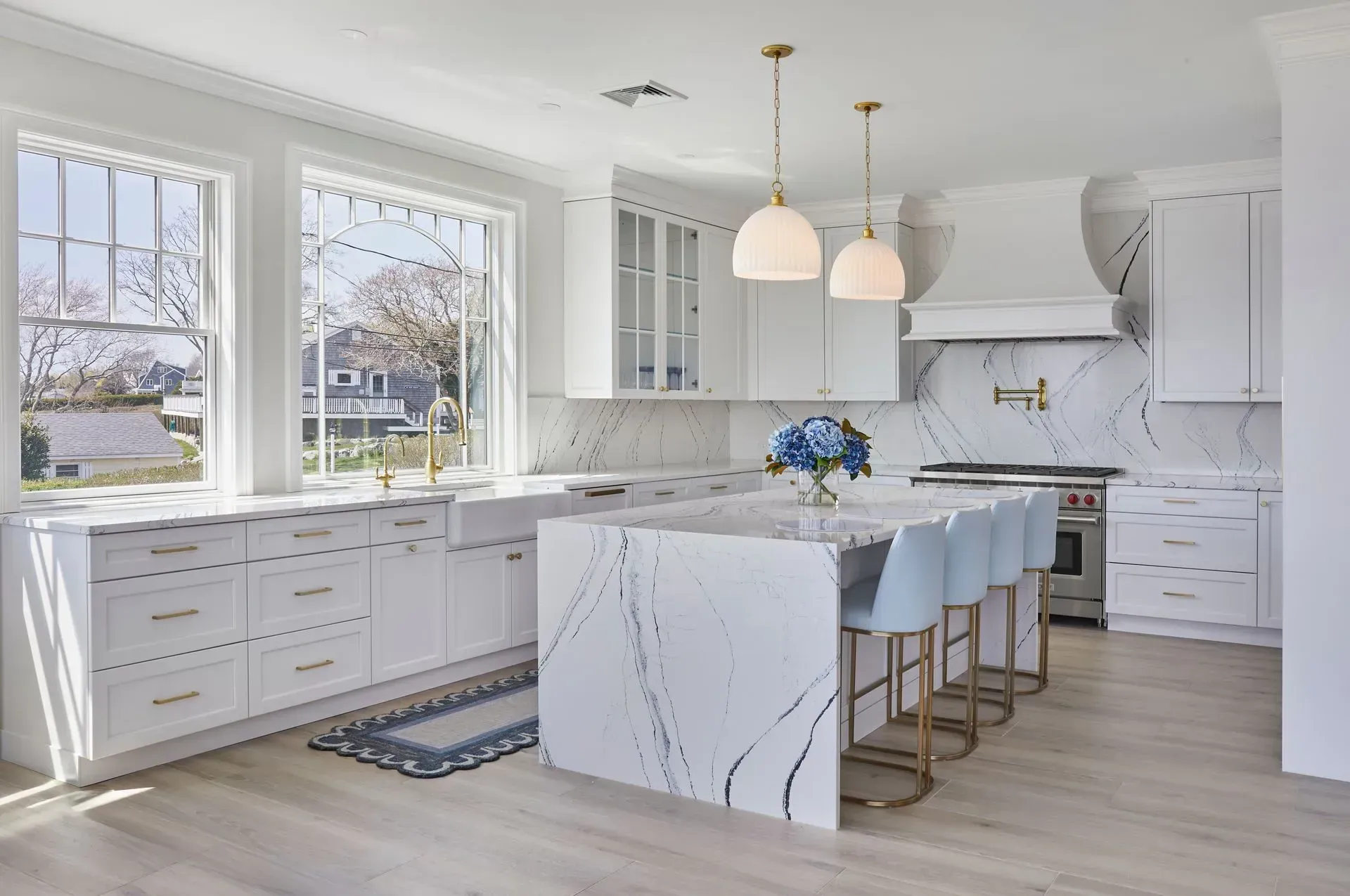 Elegant white kitchen with marble countertops, island with blue stools, and large windows.