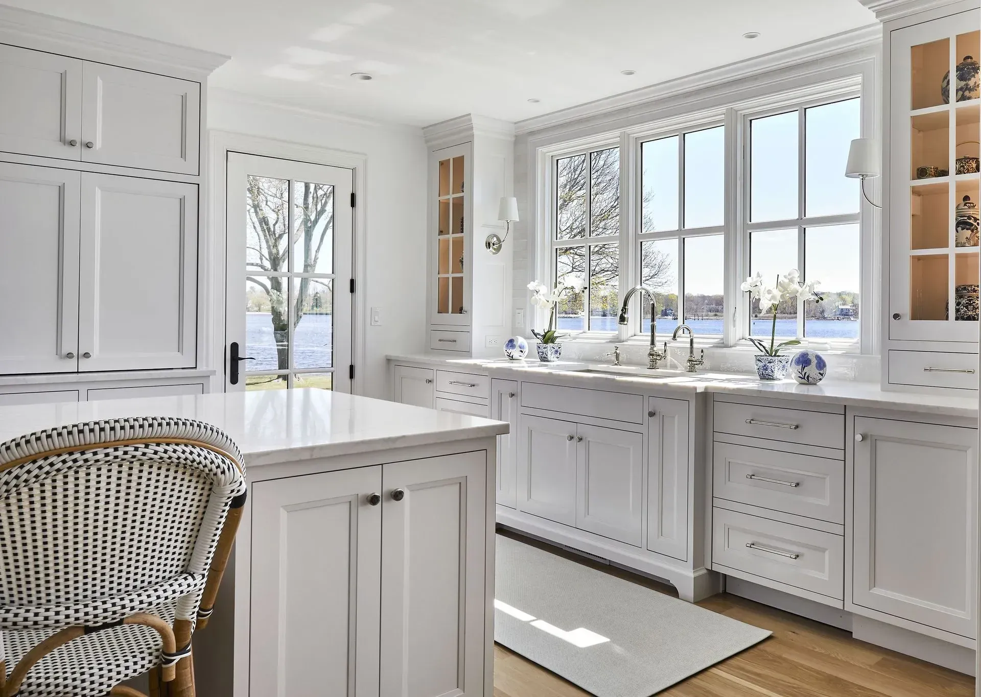 White kitchen with large window overlooking a lake. White cabinets, island, and woven chair.