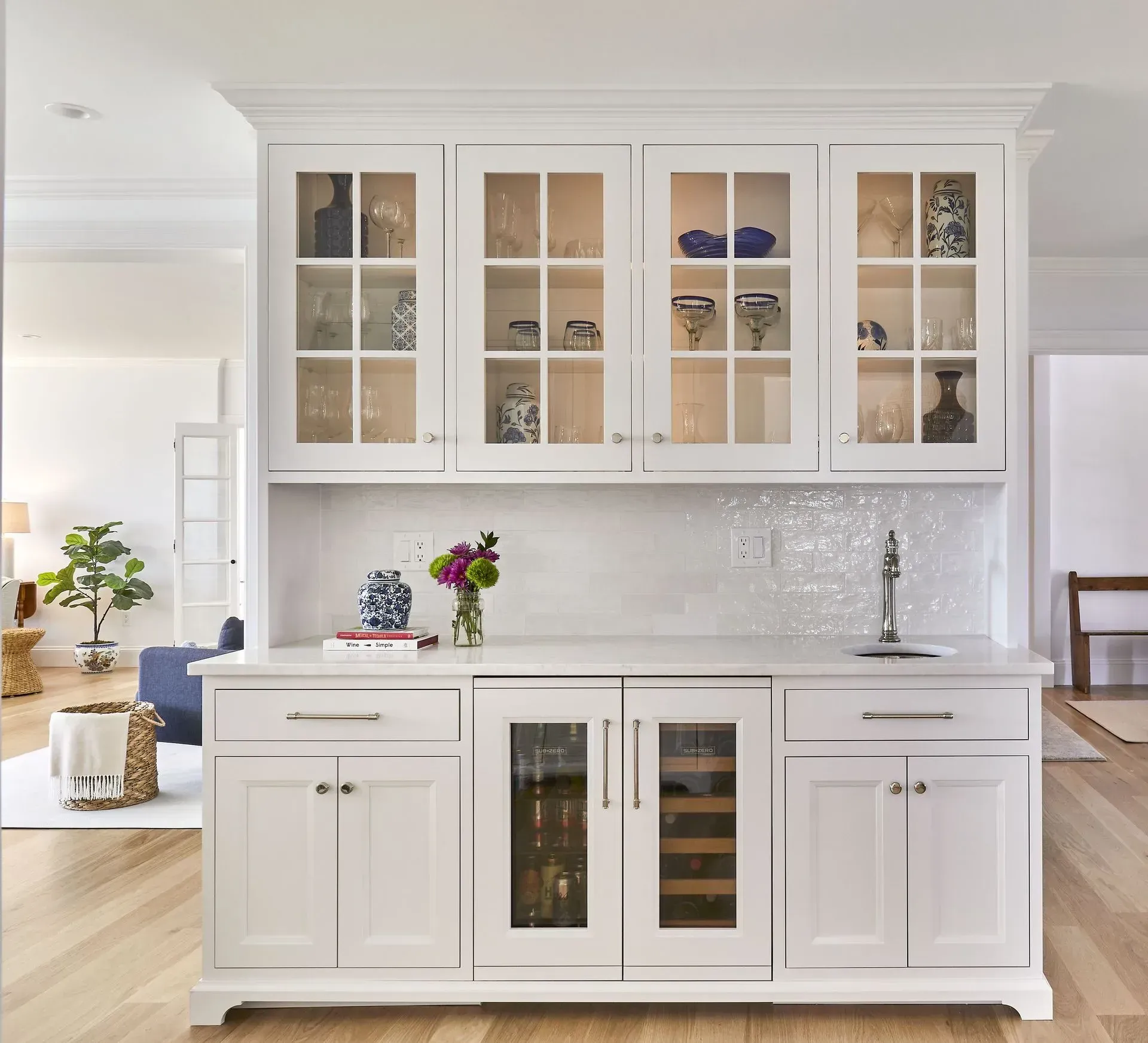 White built-in bar with glass-front cabinets, countertop, and wine cooler. Contains stemware, décor, and sink.