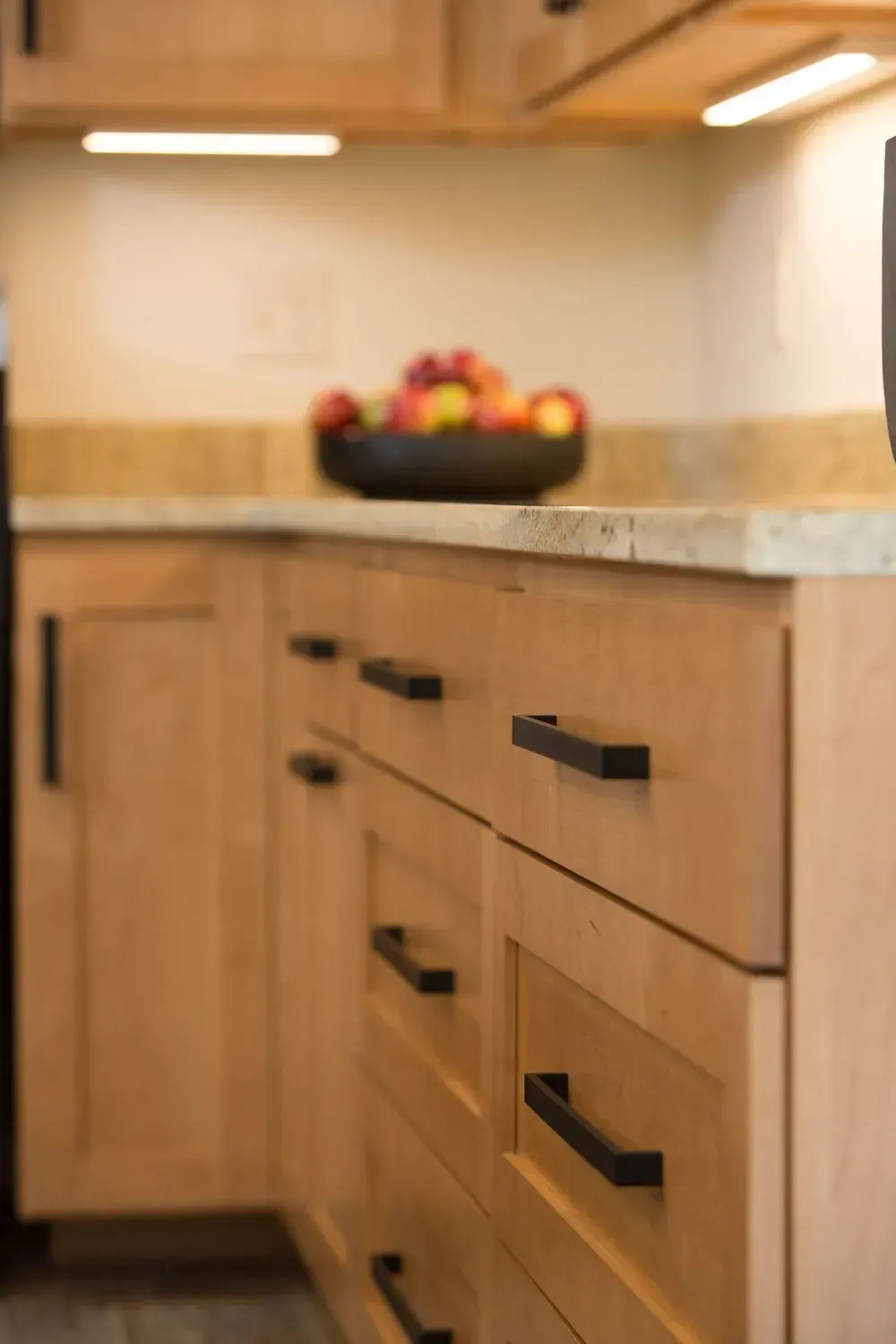 Light wood kitchen cabinets with black handles, a bowl of fruit, and a granite countertop.