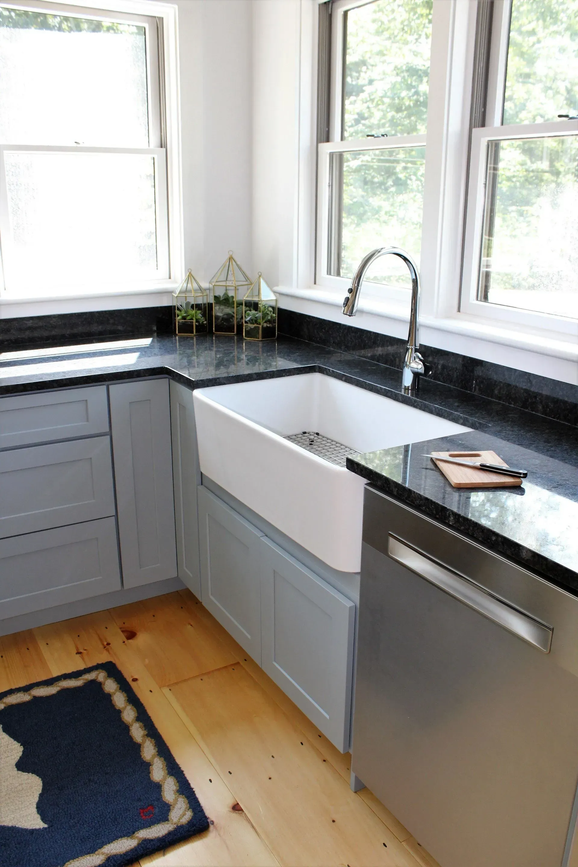 Kitchen corner with farmhouse sink, stainless dishwasher, gray cabinets, black countertops, and windows.