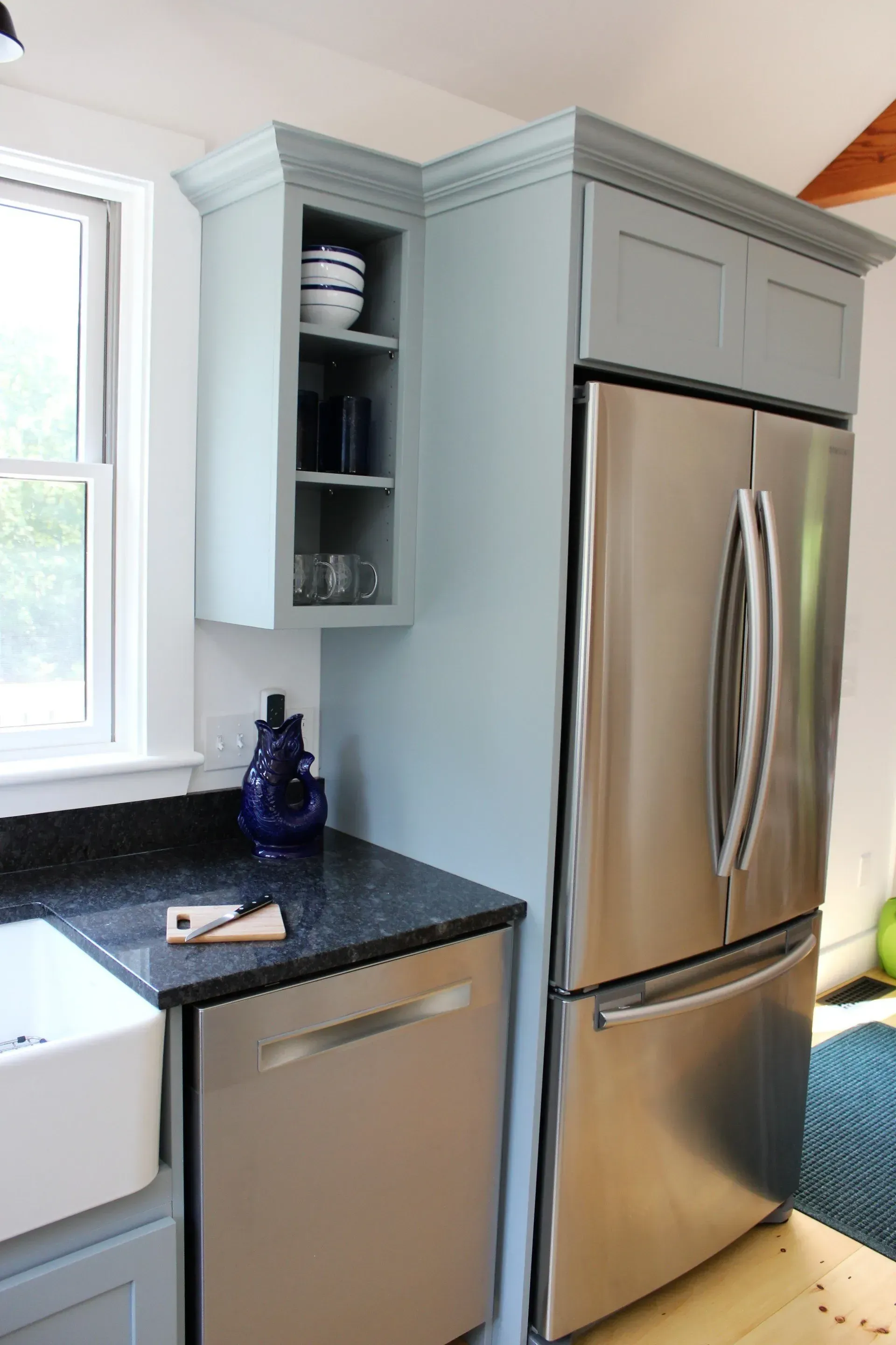 Kitchen corner with blue cabinets, stainless steel refrigerator, black countertop, white sink, and window.