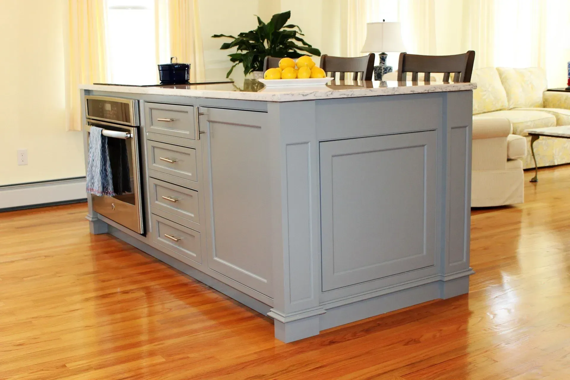 Gray kitchen island with built-in oven and drawers, hardwood floor, and a living room in the background.