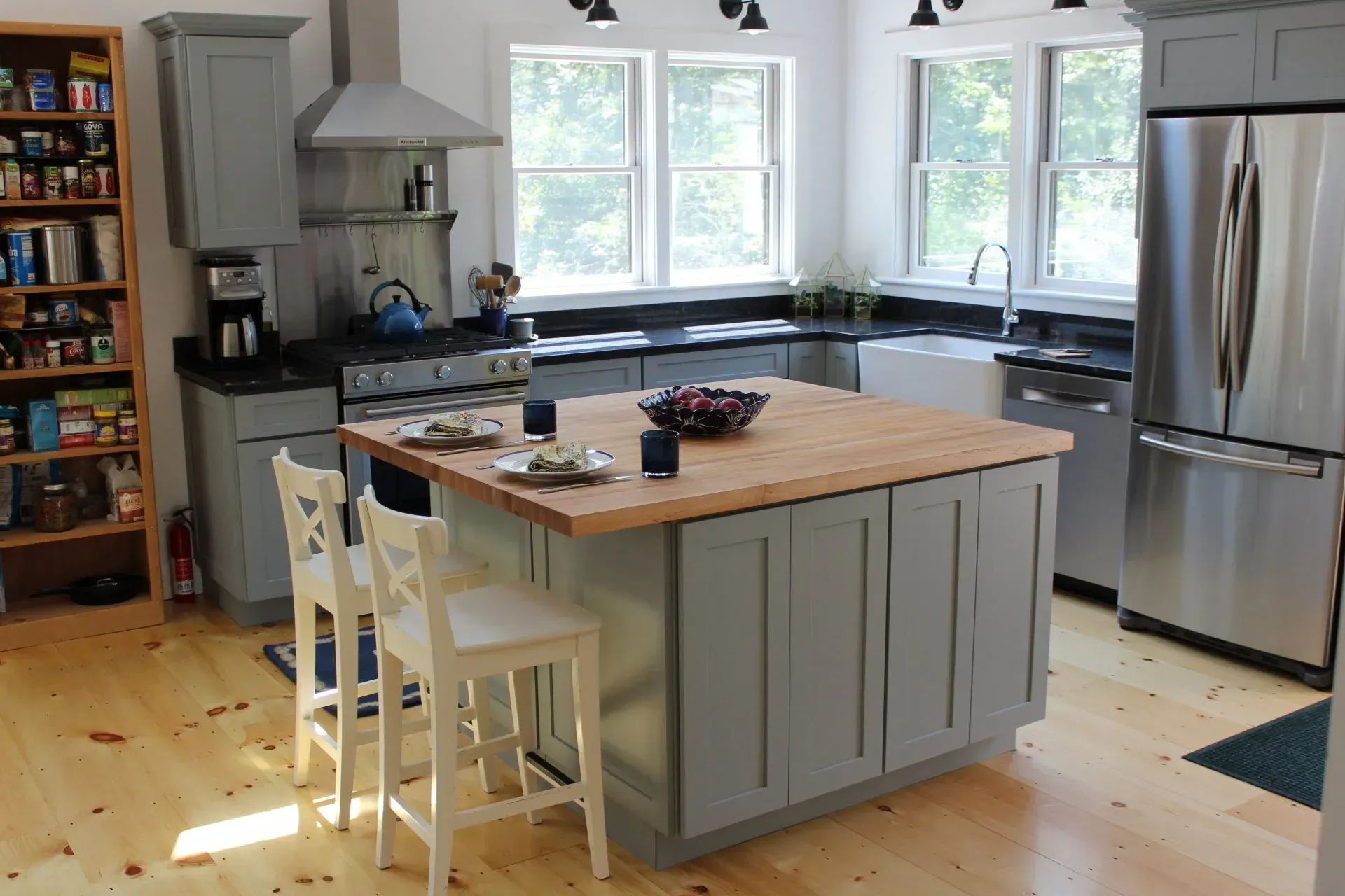 A modern kitchen with a gray island, stainless steel appliances, wood floor, and two white chairs.